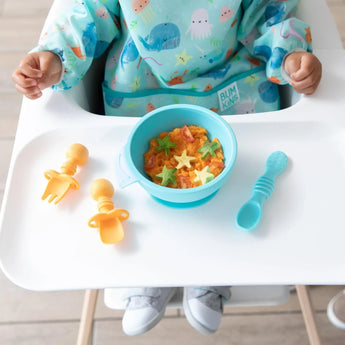 A child sits in a high chair wearing a blue bib, with a blue bowl of food, spoon, and Bumkins Silicone Chewtensils®: Tangerine on the tray.