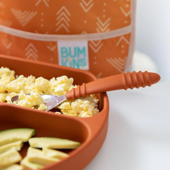 Close-up of a divided orange plate with eggs, avocado, and a Bumkins Spoon + Fork: Clay, with a blurred orange bib in the background.