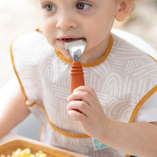 Load image into Gallery viewer, A toddler in a bib holds a Bumkins Spoon + Fork: Clay in their mouth, eating eggs from a brown plate.