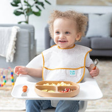Load image into Gallery viewer, A toddler in a high chair holds a Bumkins Spoon + Fork: Clay with food on a plate and toys behind.