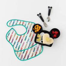 Load image into Gallery viewer, A Bumkins SuperBib® Mickey & Friends MVP bib lays flat on a white background next to a Mickey shaped plate of food and matching utensils.