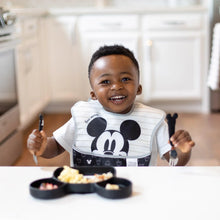 Load image into Gallery viewer, A smiling child holds Bumkins Spoon + Fork: Mickey Mouse while seated at a white table in a kitchen with a divided plate of food in front.