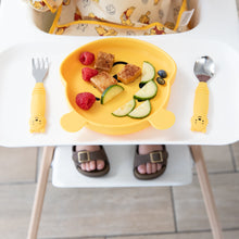 Load image into Gallery viewer, Bumkins Spoon + Fork: Winnie the Pooh sits beside a matching plate with fruit & veggies on a high chair tray; a child's feet visible below.