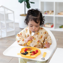 Load image into Gallery viewer, A child in a yellow patterned bib sits in a high chair, holding a Bumkins Spoon + Fork: Winnie the Pooh near a bear-shaped plate of food.