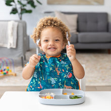Load image into Gallery viewer, A curly-haired toddler in a blue bib smiles, Bumkins Spoon + Fork: Gray above a gray divided plate on a white table in a cozy living room.