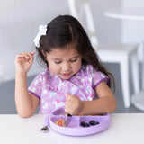 A young girl with a hair bow eats from a purple divided plate at a white table using Bumkins Spoon + Fork: Lavender, wearing a unicorn bib.