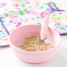 Load image into Gallery viewer, Bumkins Spoon + Fork: Pink and Bumkins Silicone Grip Bowl: Pink with cereal and milk, beside a colorful bib on a white background.