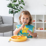 A toddler wearing a star bib uses Bumkins Spoon + Fork: Pineapple to eat from a yellow plate in a cozy living room.