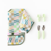Load image into Gallery viewer, Bumkins Sleeved Bib: Motley Check lays folded next to two sets of light green toddler utensils on a white background.