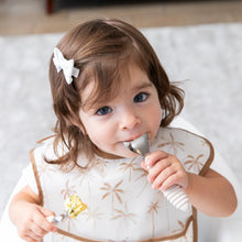 Load image into Gallery viewer, A girl wearing a beige bib holds Bumkins Spoon + Fork: Sand, one in her mouth, as she looks curiously at the camera.