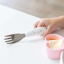Load image into Gallery viewer, Close-up of a child's hand gripping a Bumkins Spoon + Fork: Vanilla Sprinkle at a white table next to a pink heart-shaped cup of yogurt.