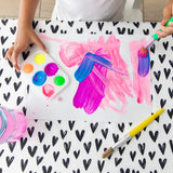 A child paints with brushes on paper at a table covered with a Bumkins Splat Mat: Hearts, with a paint palette nearby.