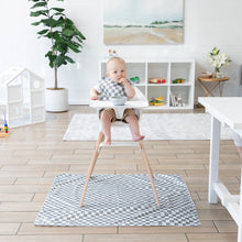 Load image into Gallery viewer, A baby in a high chair with a bib sits on a Bumkins Splat Mat: Charcoal Check, surrounded by bright modern decor.