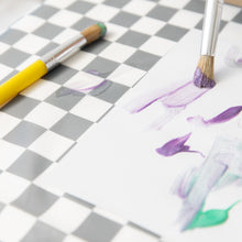 Load image into Gallery viewer, Yellow and white paint brushes sit next to a white paper with purple and green paint on top of a Bumkins Splat Mat: Charcoal Check.