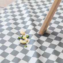 Load image into Gallery viewer, Tiny pasta and broccoli sit on a Bumkins Splat Mat: Charcoal Check, under a wooden table leg on the floor.