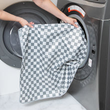 Load image into Gallery viewer, A person places Bumkins Splat Mat: Charcoal Check into an open front-loading washing machine.