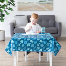 Load image into Gallery viewer, A child paints on a table covered by a Bumkins Splat Mat: Blue Tropic in front of a gray sofa and plant in a bright living room.