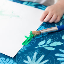 Load image into Gallery viewer, Close-up of a child's hand and a paintbrush with green paint near a white paper on a Bumkins Splat Mat: Blue Tropic.