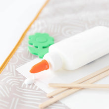 Load image into Gallery viewer, Close-up of a leaking white glue bottle with orange tip, green foam flower, wooden sticks, and white paper on a Bumkins Splat Mat: Wander.