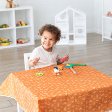 Load image into Gallery viewer, A child smiling at table with craft supplies on a table covered in a Bumkins Splat Mat: Grounded, in a bright room with toys on shelves.