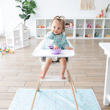 Load image into Gallery viewer, A toddler in a high chair eats from a purple bow in a playroom, with a Bumkins Splat Mat: Rainbows protecting the floor below.