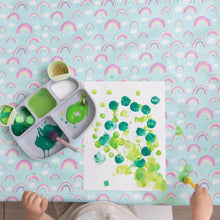Load image into Gallery viewer, On a Bumkins Splat Mat: Rainbows, a child paints green dots on paper with a silicone plate of paints and art supplies nearby.