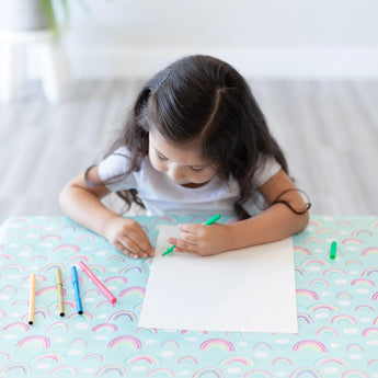 A girl draws on white paper with colored pens at a table covered by a Bumkins Splat Mat: Rainbows.
