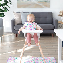 Load image into Gallery viewer, A smiling baby sits in a high chair with a colorful bib on a Bumkins Splat Mat: Watercolor, near a gray sofa and green plant.