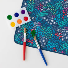 Load image into Gallery viewer, Two paintbrushes with red and blue handles sit on a Bumkins Splat Mat: Animal Prints near a palette of paints on a white background.