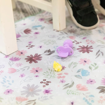 Bumkins Splat Mat: Floral lays on a floor under a chair, with a purple baby spoon and yellow food on top.