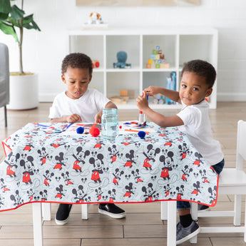 Two young children enjoy arts and crafts at a table covered by Bumkins Splat Mat: Mickey Mouse in a bright playroom.