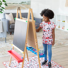 Load image into Gallery viewer, Bumkins Splat Mat: Minnie Mouse lays on a wood floor under an easel as a girl in a matching apron paints.