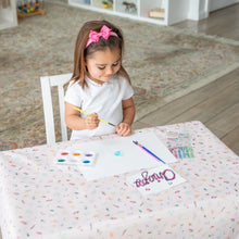 Load image into Gallery viewer, A girl with a pink bow paints with watercolors and brushes at a table with Bumkins Splat Mat: Princess Magic in a bright room.