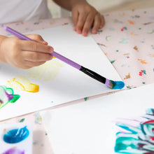 Load image into Gallery viewer, Close-up of a child painting with a purple brush on white paper on a Bumkins Splat Mat: Princess Magic.