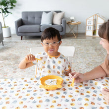Load image into Gallery viewer, A child with glasses smiles at a table covered with a Bumkins Splat Mat: Winnie and Friends, eating from a plate with an adult nearby.