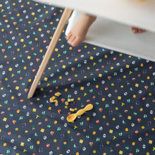 Load image into Gallery viewer, Bumkins Splat Mat: Super Mario™ Lineup lays under a high chair with a child's feet visible; a yellow spoon & food pieces scattered on top.