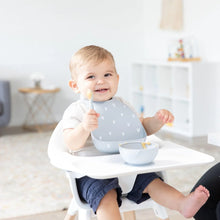 Load image into Gallery viewer, A happy baby in a high chair wears a Bumkins Silicone Bib: Hearts, holding a spoon by a gray food bowl in a bright room.