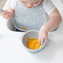 Load image into Gallery viewer, A baby in a high chair, wearing a Bumkins Silicone Bib: Hearts, plays with orange puree in a gray bowl.