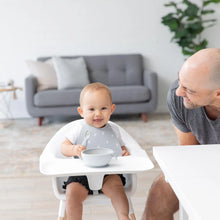 Load image into Gallery viewer, A baby in a high chair smiles with a spoon, wearing a Bumkins Silicone Bib: Hearts as an adult leans over; a gray sofa and plant are behind.