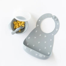 Load image into Gallery viewer, Bumkins Silicone Bib: Hearts lays next to a gray bowl & spoon set with scrambled eggs rests on a white background.