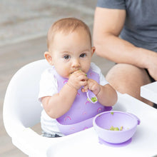 Load image into Gallery viewer, A baby in a high chair wears a Bumkins Silicone Bib: Rainbows, eating with a spoon from a purple bowl.
