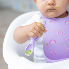 Load image into Gallery viewer, Close-up of a baby in a high chair, wearing a Bumkins Silicone Bib: Rainbows, holding a purple spoon with food-smeared hands.