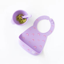 Load image into Gallery viewer, Bumkins Silicone Bib: Rainbows lays next to a purple bowl with mashed avocado and a purple spoon on a white background.