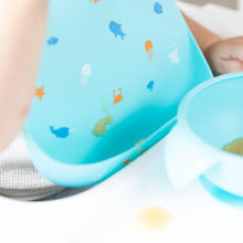 Load image into Gallery viewer, Close-up of a baby in a high chair wearing a Bumkins Silicone Bib: Ocean Life, with focus on a blob of applesauce near the front pocket.