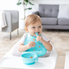 Load image into Gallery viewer, A toddler in a high chair, wearing a Bumkins Silicone Bib: Ocean Life, smiles with spoon in hand with a blue bowl nearby.