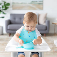 Load image into Gallery viewer, A baby in a high chair enjoys a meal from a blue bowl in a bright room, wearing a Bumkins Silicone Bib: Ocean Life.