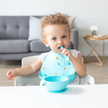 Load image into Gallery viewer, A toddler with curly hair in a high chair wearing a Bumkins Silicone Bib: Ocean Life enjoys self-feeding from a blue bowl with a spoon.