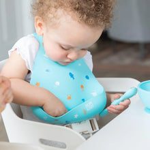 Load image into Gallery viewer, A toddler in a high chair wearing a Bumkins Silicone Bib: Ocean Life reaches into the pocket as they hold a blue spoon.
