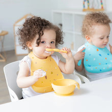 Load image into Gallery viewer, Two toddlers enjoy mealtime at a white table; one wears a Bumkins Silicone Bib: Camp Gear and eats from a yellow bowl with a spoon.