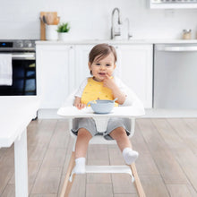 Load image into Gallery viewer, A baby in a high chair wears a Bumkins Silicone Bib: Camp Gear, smiling with a gray bowl on the tray in a modern kitchen.